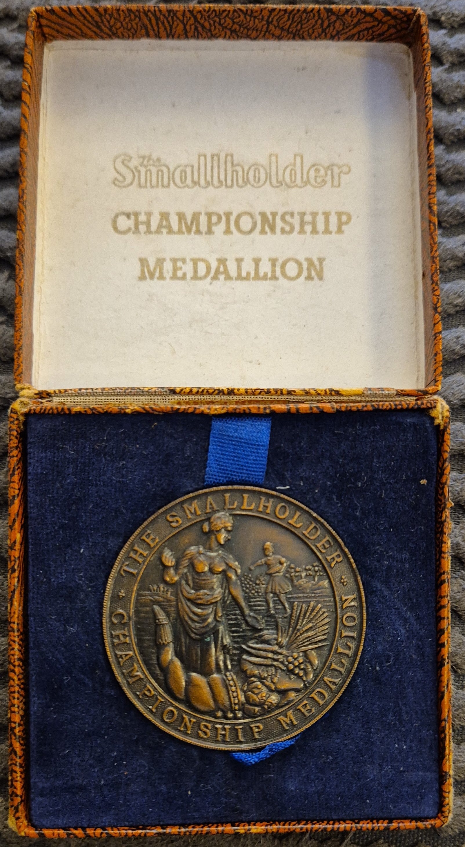 A round bronze-colored commemorative medallion displayed inside an open presentation box with a blue velvet insert and gold-toned trim. The inside lid reads 'Smallholder CHAMPIONSHIP MEDALLION.